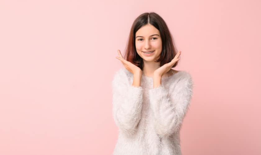 Woman in white sweater smiling with hands near face against pink background